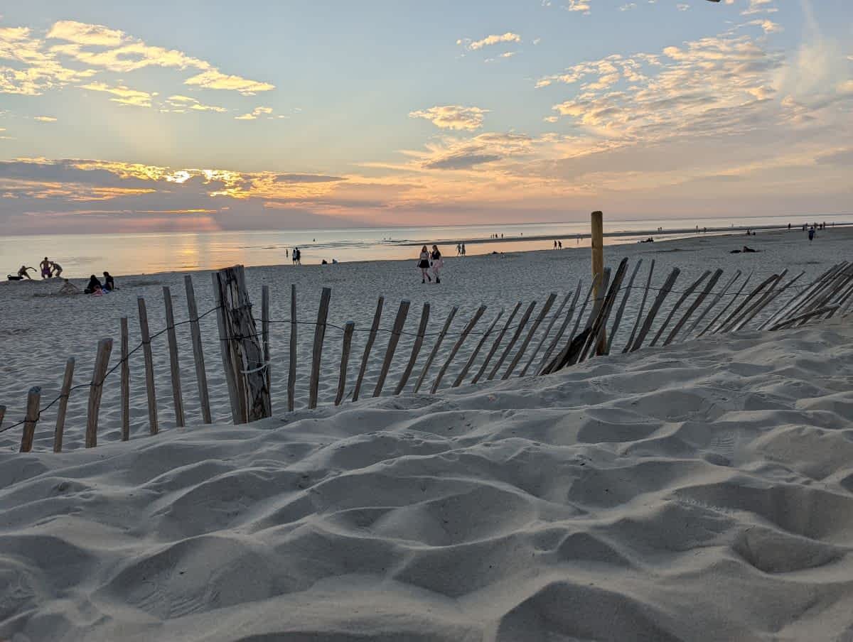 Blick über den Sand und einen verwitterten Holzzaun in den Dünen auf den weiten Nordseestrand bei Sonnenuntergang.