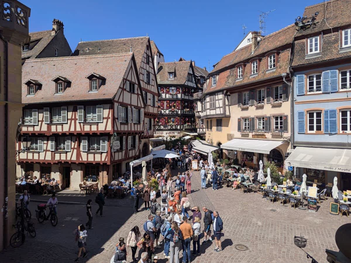 Belebter Platz in der Altstadt von Colmar mit zahlreichen Touristen, Cafés und prächtigen, blumengeschmückten Fachwerkhäusern unter blauem Himmel.
