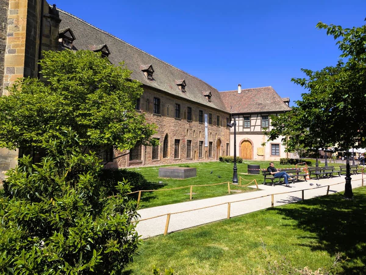 as historische Gebäude des Unterlinden-Museums in Colmar mit grünem Vorplatz und einer Parkbank im Sonnenschein.