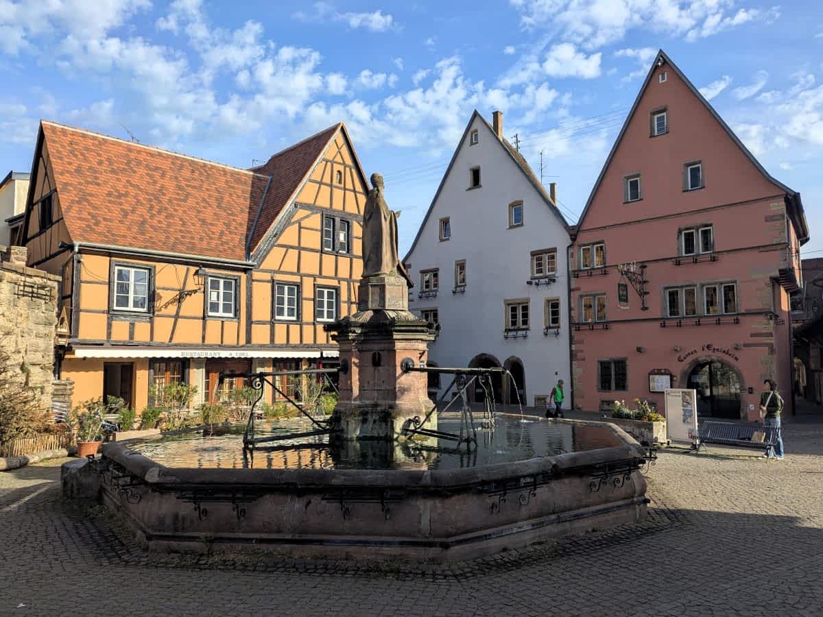 Der achteckige St.-Leo-Brunnen auf dem Marktplatz von Eguisheim vor farbenfrohen Fachwerkhäusern unter blauem Himmel.