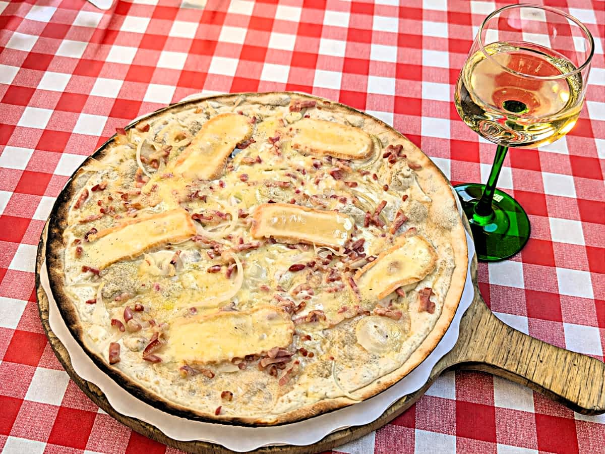 Ein elsässischer Flammkuchen mit Munsterkäse auf einem Holzbrett, daneben ein Glas Weißwein auf einer rot-weiß-karierten Tischdecke.