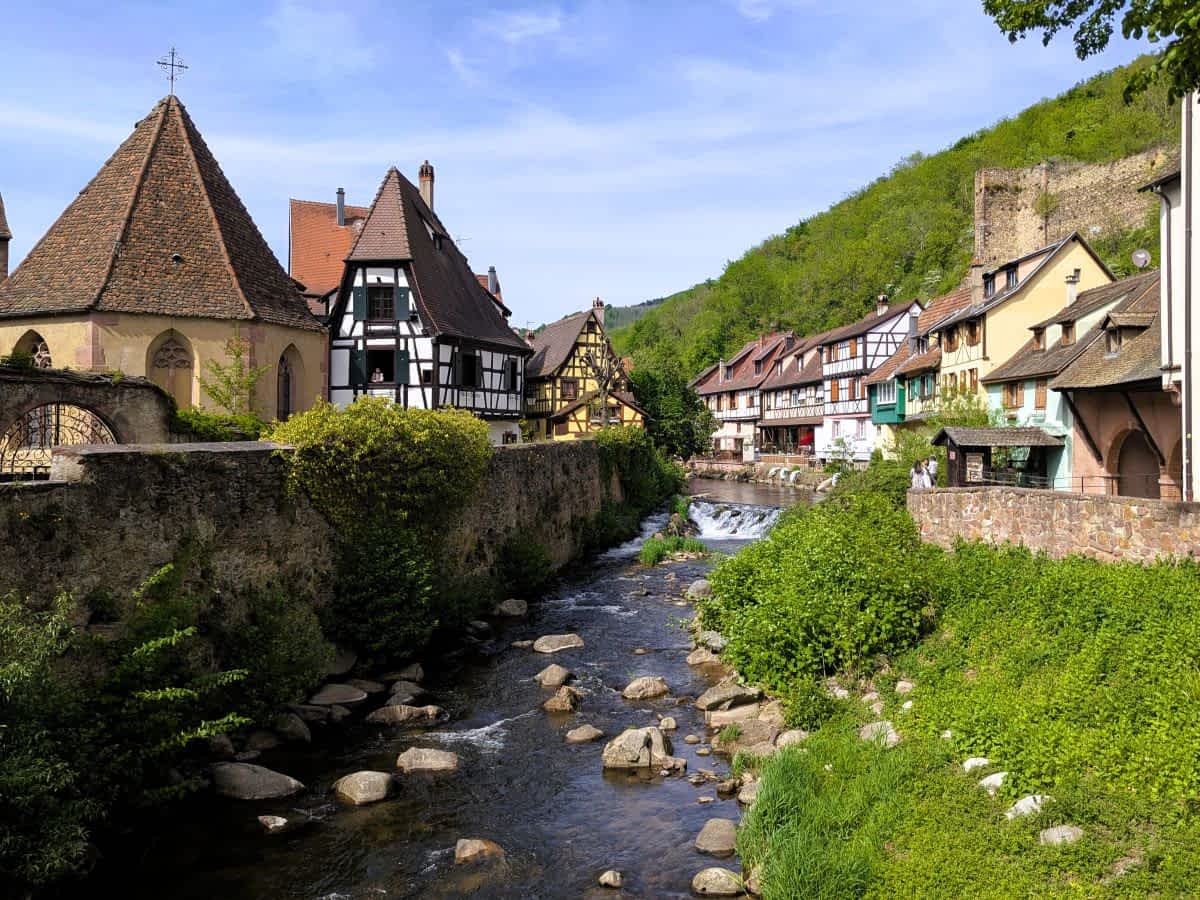 lick über den Fluss Weiss auf die historischen Fachwerkhäuser von Kaysersberg mit der Burgruine im Hintergrund unter blauem Himmel.
