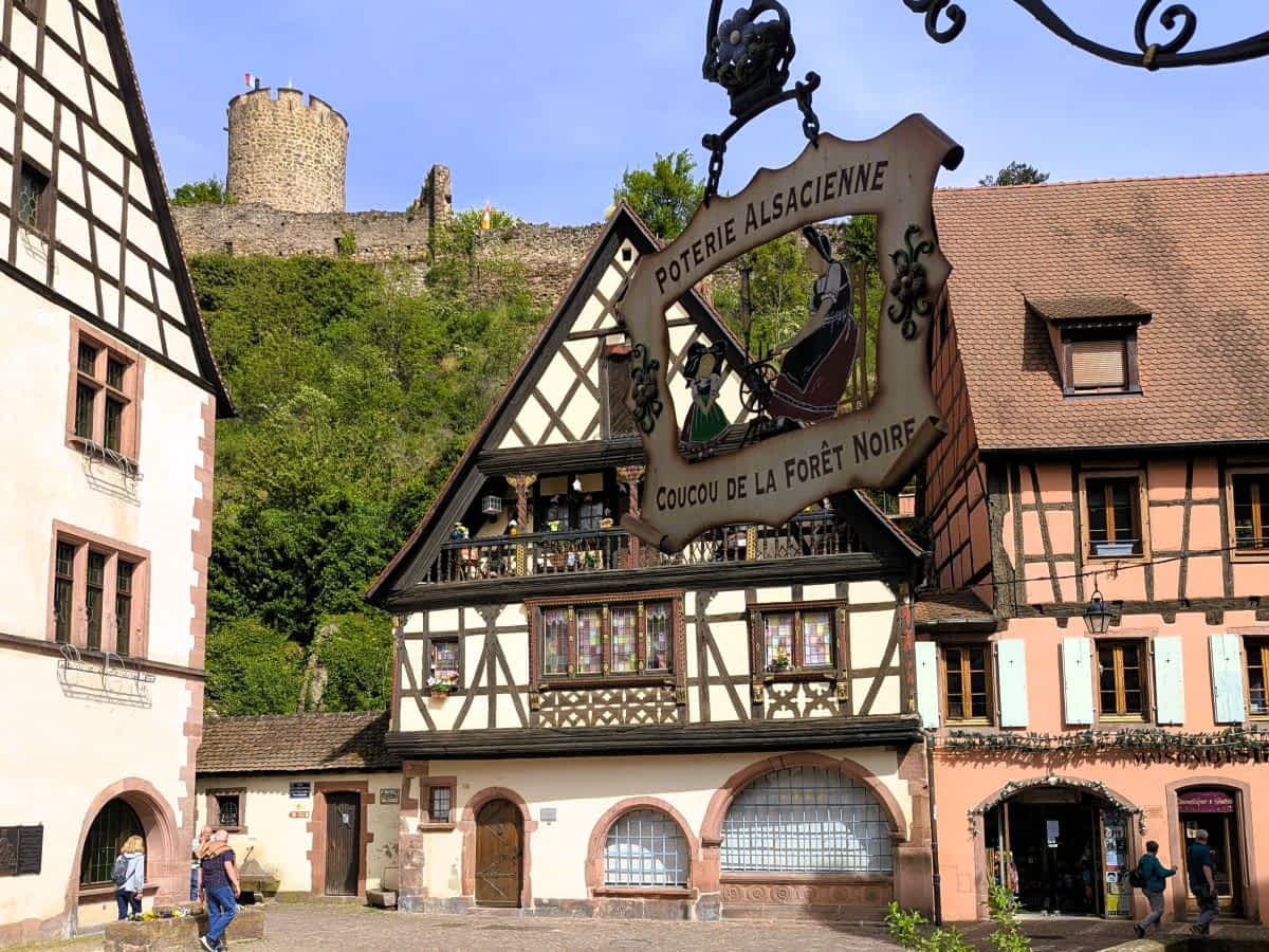 Historischer Platz in Kaysersberg mit prächtigen Fachwerkhäusern und der mittelalterlichen Burgruine auf dem bewaldeten Hügel im Hintergrund.