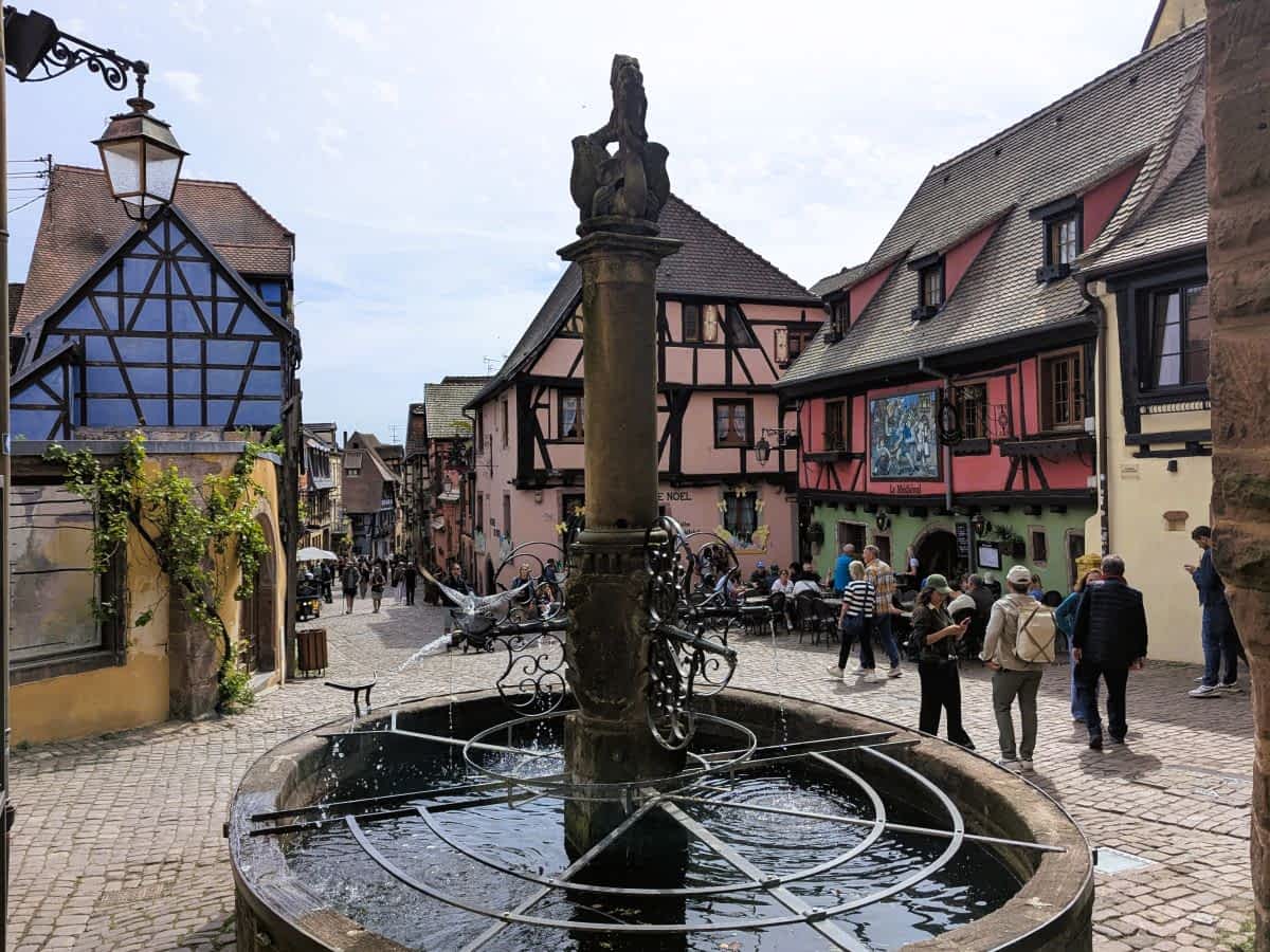 Ein historischer Steinbrunnen auf dem Place de la Sinn in Riquewihr, umgeben von Touristen und bunten Fachwerkhäusern unter einem hellen Himmel.