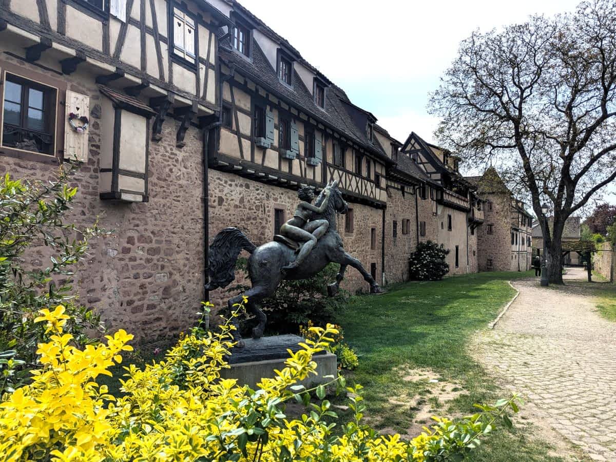 Historische Stadtmauer von Riquewihr mit aufgesetzten Fachwerkhäusern, einer Bronzestatue eines Reiters im Vordergrund und gelben Blumen.
