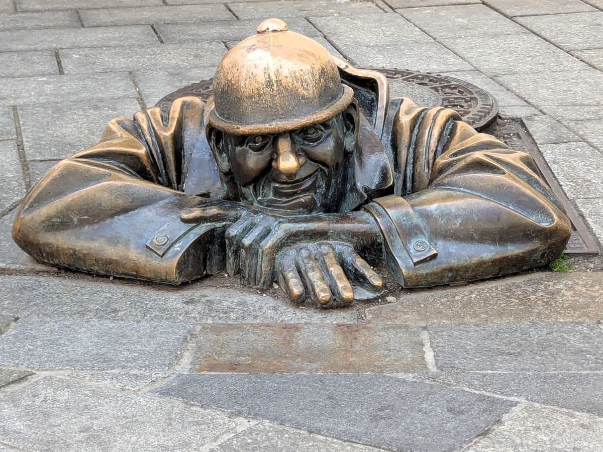 Nahaufnahme der bekannten Bronzestatue „Čumil“ (der Gucker) in Bratislava, die einen Arbeiter darstellt, der aus einem offenen Kanalschacht auf dem gepflasterten Bürgersteig herausblickt.
