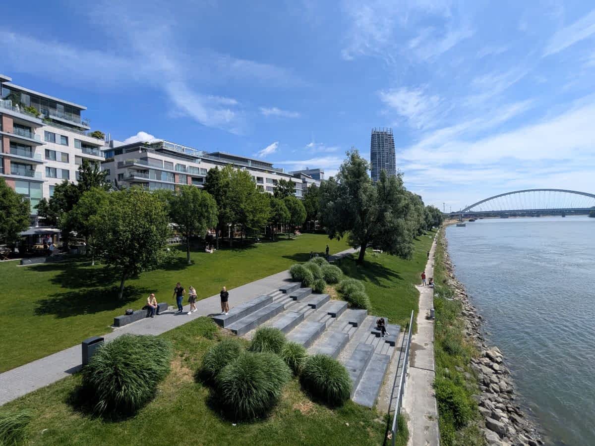 Blick entlang der modernen Donaupromenade in Bratislava. Grüne Rasenflächen und Baumreihen säumen das Flussufer. Menschen sitzen auf steinernen Stufenanlagen oder spazieren. Rechts fließt die Donau, im Hintergrund ist eine moderne Brücke und ein Hochhaus zu sehen.