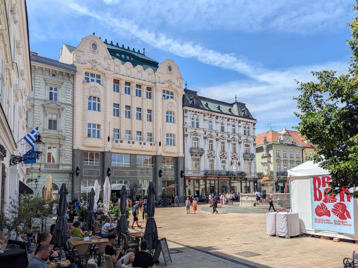 Der sonnige Hauptplatz (Hlavné námestie) in Bratislava mit historischen Gebäuden im Hintergrund, darunter prachtvolle Fassaden und ein Brunnen. Im Vordergrund sind Tische eines Straßencafés und ein weißes Zelt des Bratislavaer Buchfestivals zu sehen.