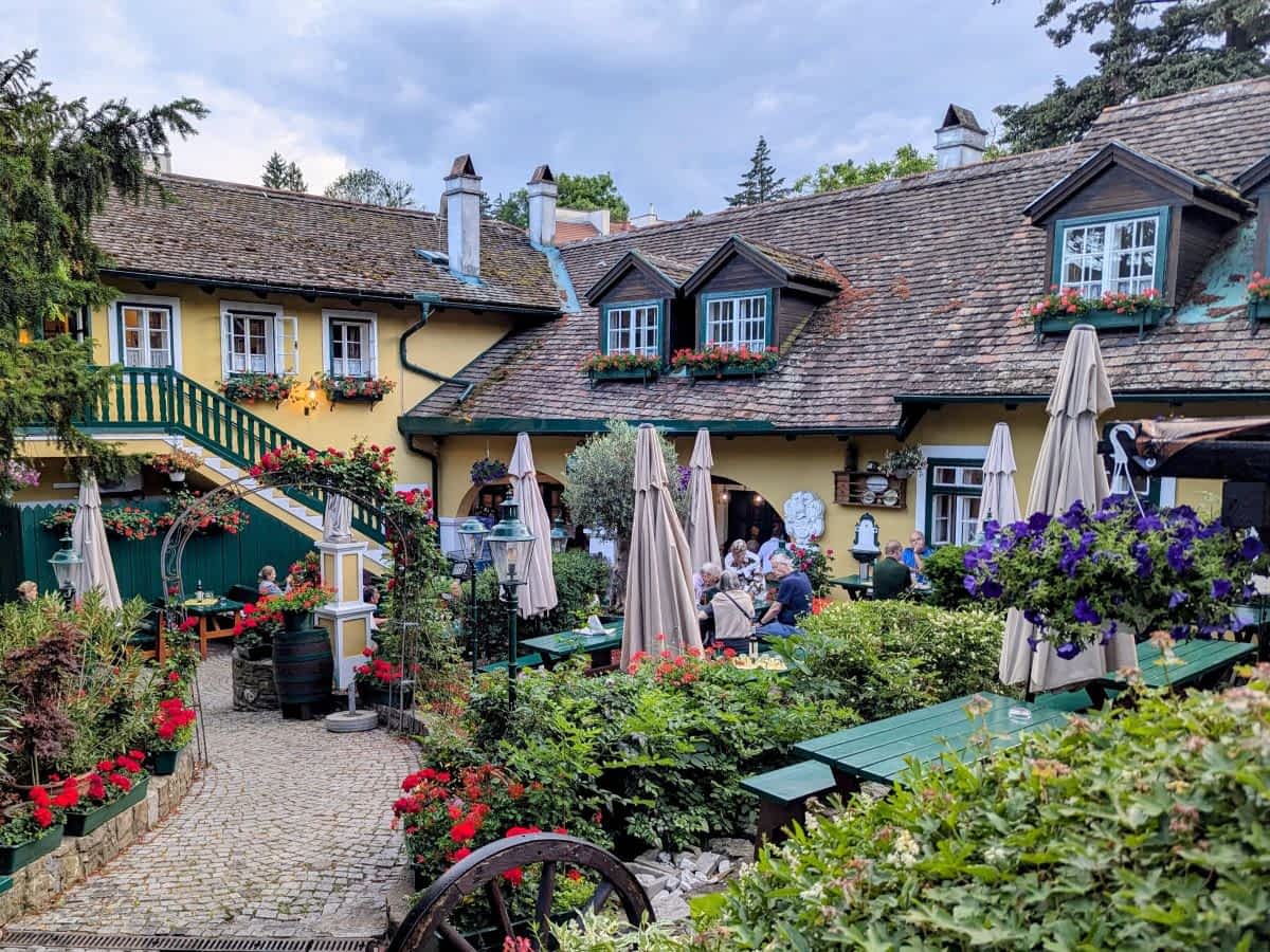 Idyllischer Heuriger in Wien mit Gastgarten, Blumenbeeten, Holzbänken und typischer Weinlokal-Atmosphäre im Grünen.
